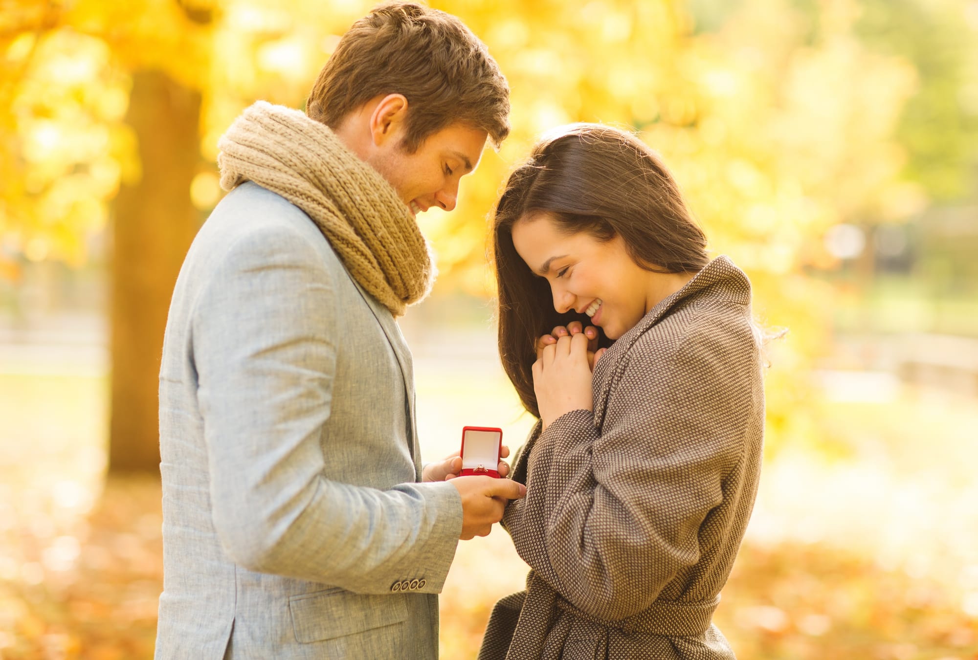 A happy couple under autumn trees. The man is proposing, presenting the woman with a red engagement ring box.